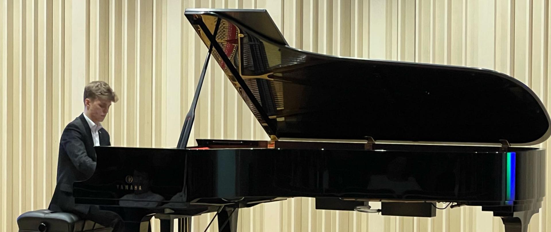 Na zdjęciu chłopak w białej koszuli i czarnym garniturze, który siedzi na czarnej ławie i gra na czarnym fortepianie. Tło obrazka w odcieniach beżu. Zdjęcie zostało wykonane podczas koncertu fortepianowego. 