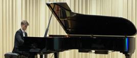 Na zdjęciu chłopak w białej koszuli i czarnym garniturze, który siedzi na czarnej ławie i gra na czarnym fortepianie. Tło obrazka w odcieniach beżu. Zdjęcie zostało wykonane podczas koncertu fortepianowego. 