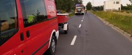 Dwa pojazdy pożarnicze, jedne z podczepioną łodzią stoją na drodze wzdłuż lasu.