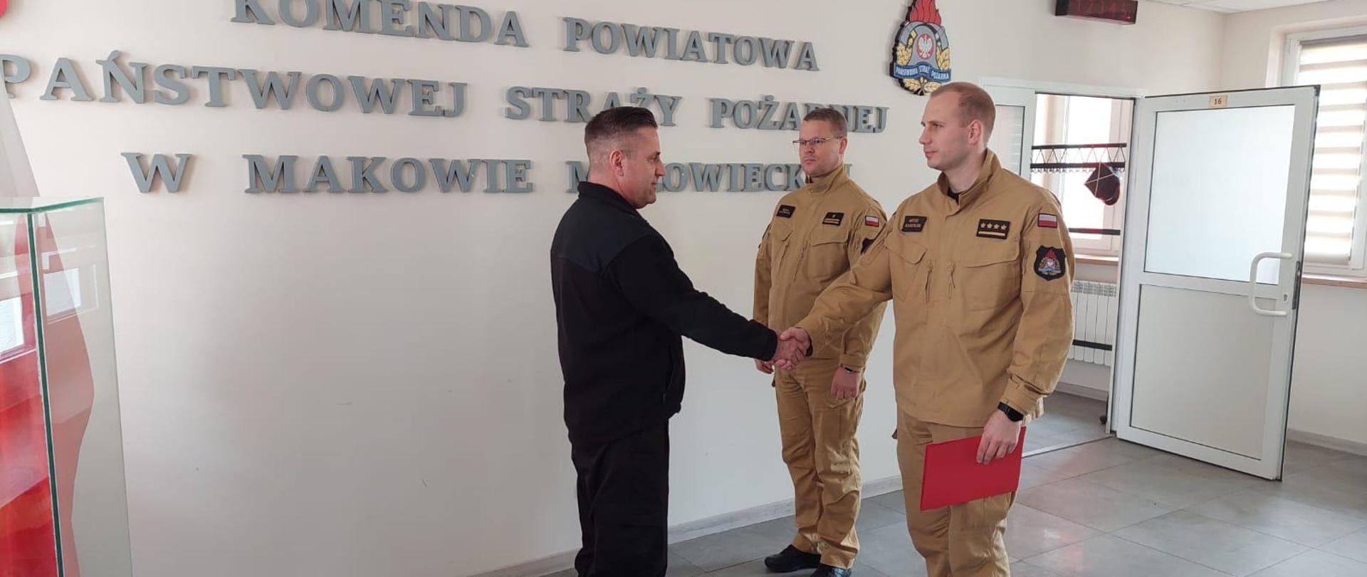 Komendant składa gratulacje funkcjonariuszowi poprzez uścisk ręki, w drugim ręku trzyma czerwoną teczkę. Obok niego stoi funkcjonariusz.