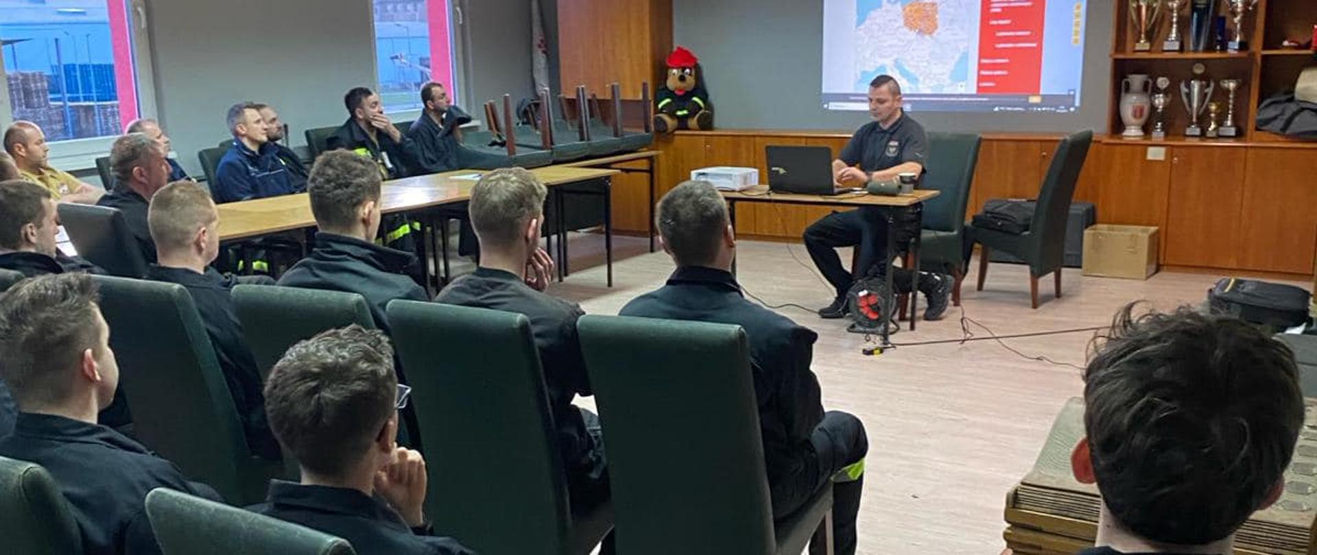 Na zdjęciu znajdują się strażacy ubrani w umundurowanie koszarowe, siedzący na sali wykladowej