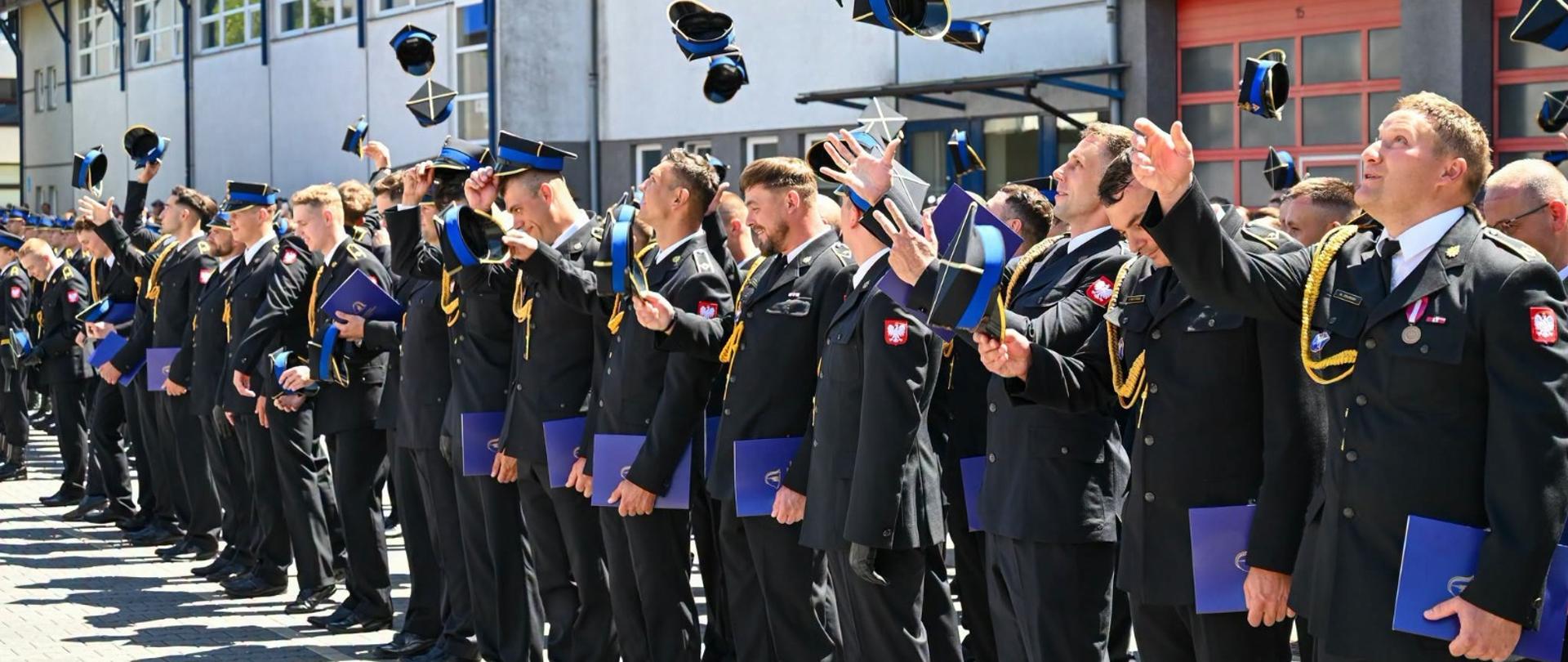 Grupa strażaków Państwowej Straży Pożarnej w galowych mundurach stoi w szeregu na placu przed budynkiem komendy. Wszyscy z uśmiechem i radością podrzucają w górę swoje czapki rogatywki podczas uroczystego apelu. W rękach trzymają niebieskie foldery lub dyplomy. W prawym dolnym rogu widnieje podpis: „st. str. kdt Piotr Krzak | Państwowa Straż Pożarna”.