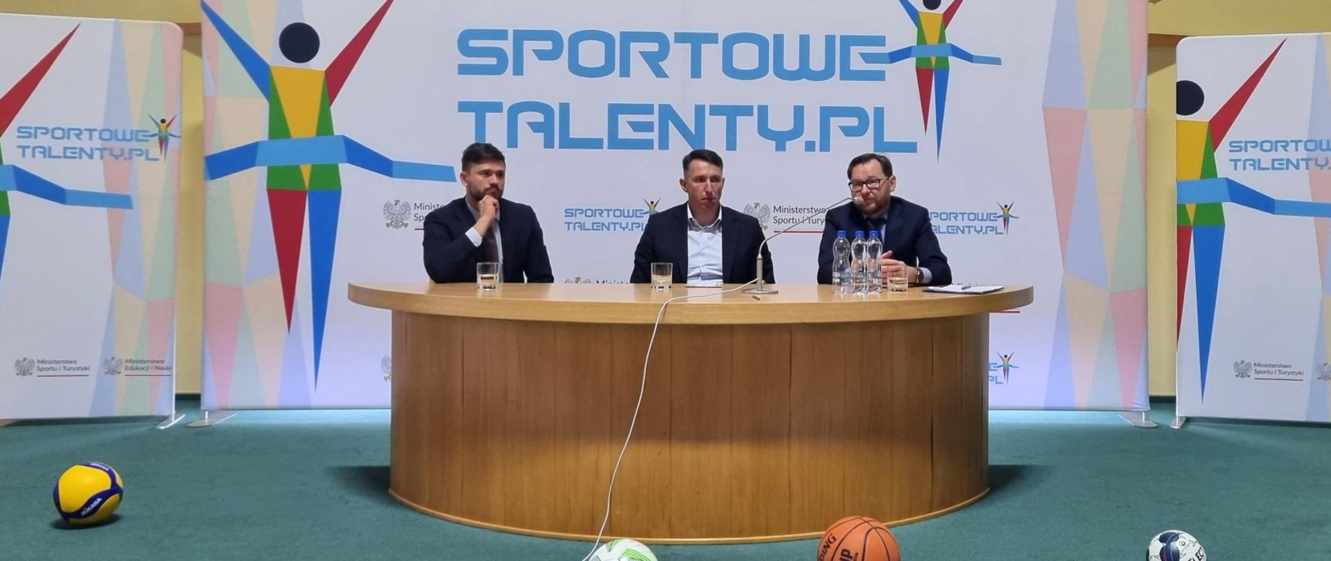 Na zdjęciu znajduje się trzech prelegentów spotkania z dyrektorami szkół woj. opolskiego dot. pilotażowej edycji programu Sportowe Talenty. Prelegenci siedzą za półowalnym stołem. W tle znajdują się materiały wizerunkowe Sportowych Talentów.
