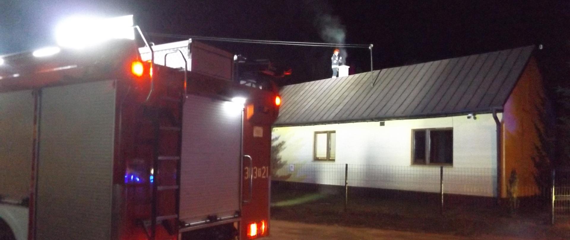 Zdjęcie przedstawia samochód gaśniczy stojący przed posesją, na której doszło do pożaru sadzy. W tle, na dachu budynku widać strażaka stojącego przy kominie. Z Komina wydobywa się dym. 