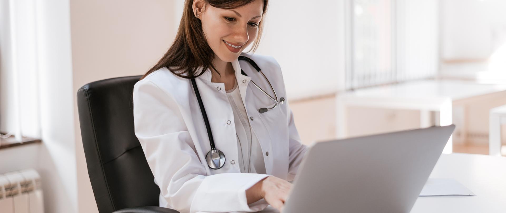 Młoda, uśmiechnięta lekarka pracuje na laptopie.