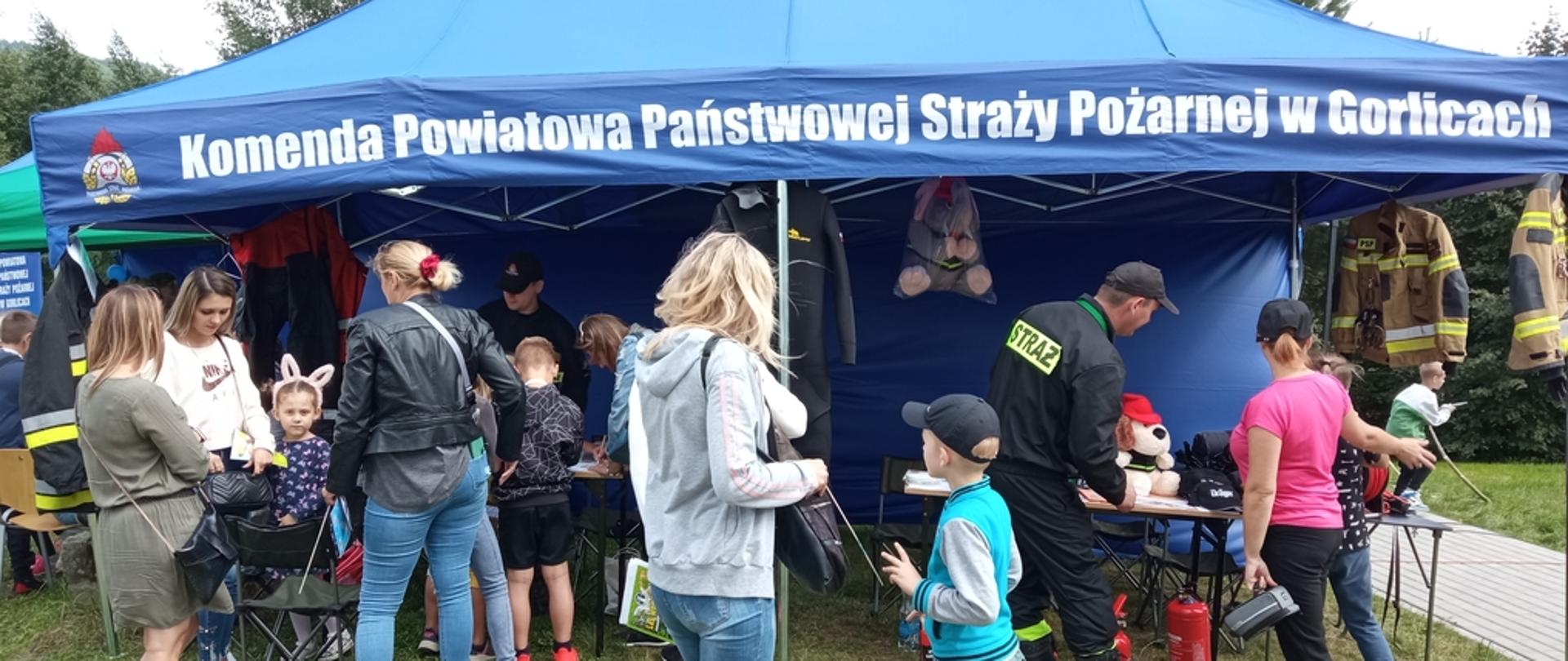 Na zdjęciu widoczna grupa dzieci i osób dorosłych uczestniczących w pikniku rodzinnym przed namiotem koloru niebieskiego z napisem Komenda Powiatowa PSP w Gorlicach pod którym strażacy prowadzili prelekcje i konkursy z zakresu bezpieczeństwa przeciwpożarowego.
