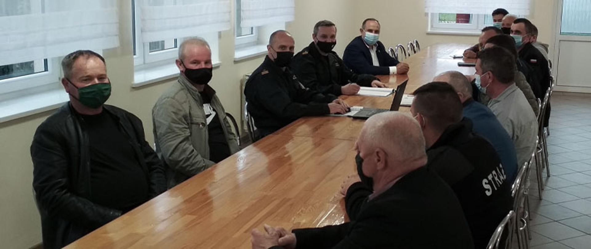 Zdjęcie przedstawia piętnaście osób siedzących przy długim drewnianym stole po obu stronach, po lewej stronie stołu siedzi komendant powiatowy PSP w Łowiczu mł. bryg. Arkadiusz Makowski oraz jego zastępca mł. bryg. Rafał Garbarz, po lewej ręce zastępcy komendanta siedzi wójt gminy Łyszkowice Adam Ruta, po przeciwnej stronie stołu siedzą druhowie oraz po ich środku wiceprzewodniczący Rady Powiatu Łowickiego Waldemar Krajewski. Wszystkie osoby mają maski chirurgiczne na twarzy. Pomieszczenie z terakotą na podłodze, po lewej stronie stołu znajdują się okna z roletami, w szczycie stołu na wprost jedno okno z roletą i drzwi.