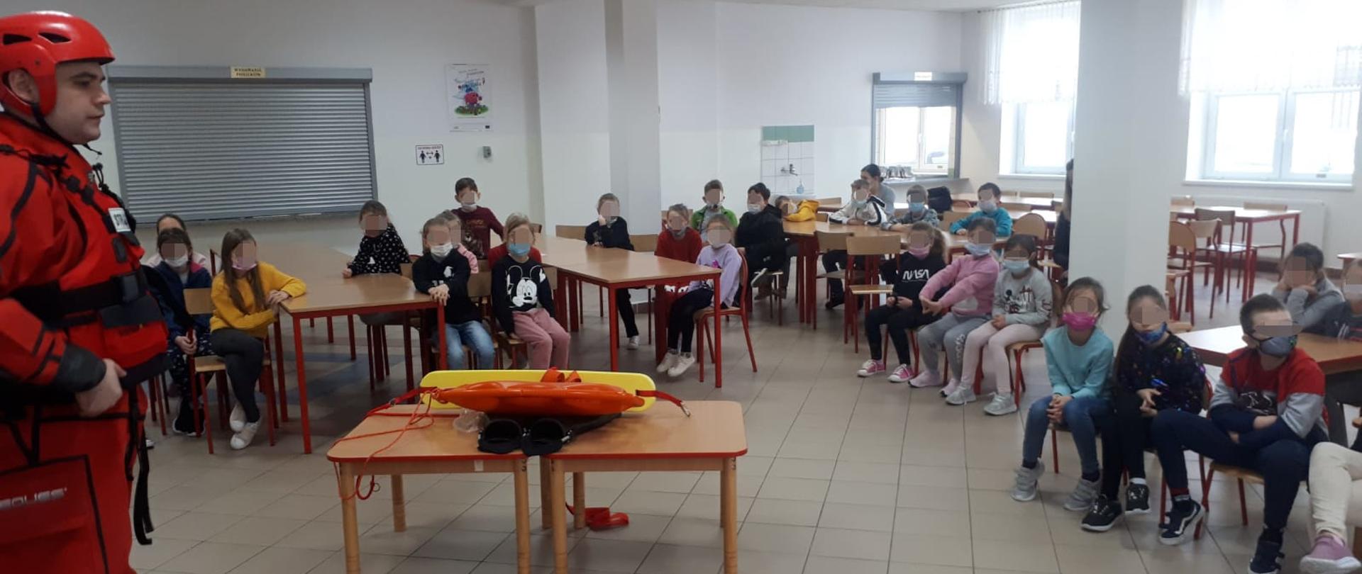 Zdjęcie wykonane jest na świetlicy i przedstawia strażaka ubranego w ubranie wypornościowe do pracy w wodzie i kask oraz siedzących na krzesełkach i słuchających go dzieci. Na stoliku między dziećmi i strażakiem znajduje się sprzęt wykorzystywany w ratownictwie wodno-lodowym. Po prawej stronie zdjęcia widać również manekiny do udzielania pierwszej pomocy. 