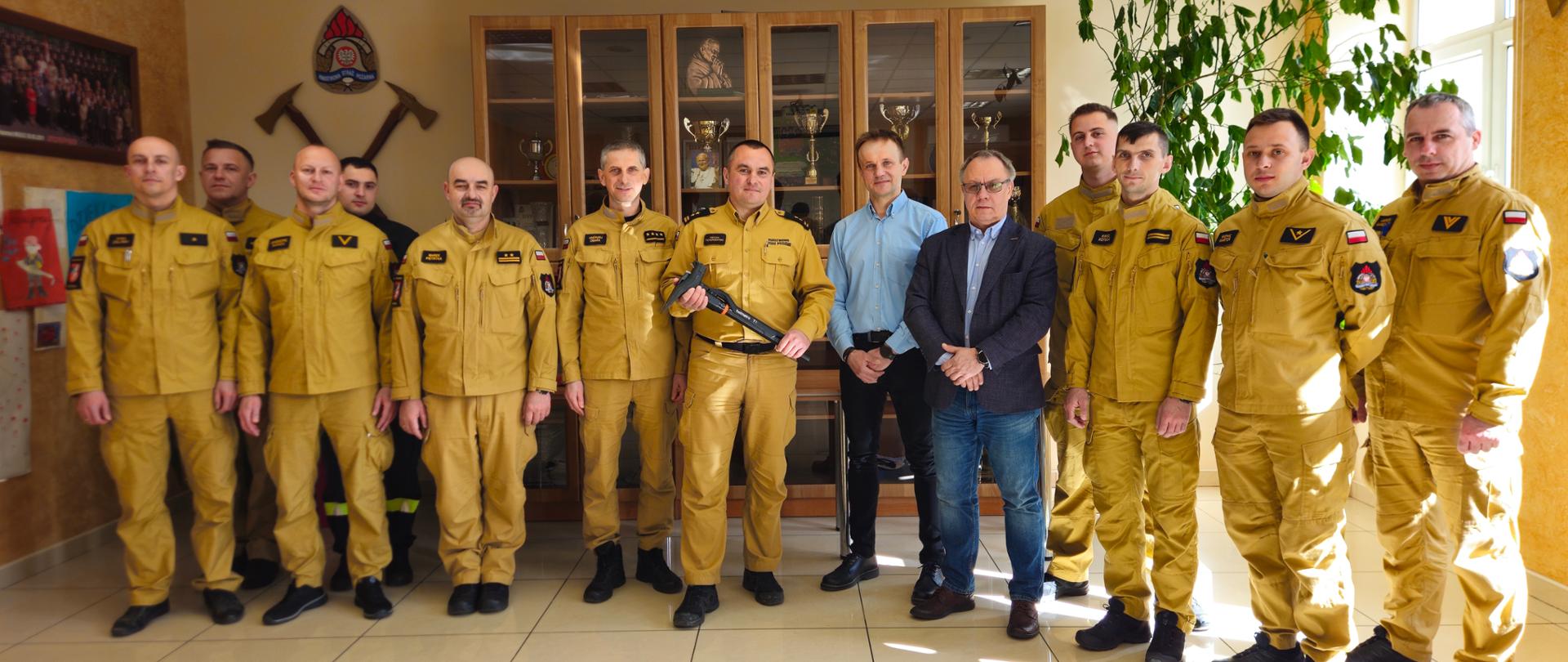 Grupa strażaków ubranych w jasnożółte mundury strażackie oraz dwóch mężczyzn w ubraniach cywilnych stoi we wnętrzu budynku Komendy Powiatowej PSP w Mielcu. Na środku, jeden z funkcjonariuszy trzyma w dłoniach wielofunkcyjne narzędzie do wejść siłowych T1 marki Holmatro. W tle widać przeszkloną gablotę z pucharami oraz symbole strażackie zawieszone na ścianie.