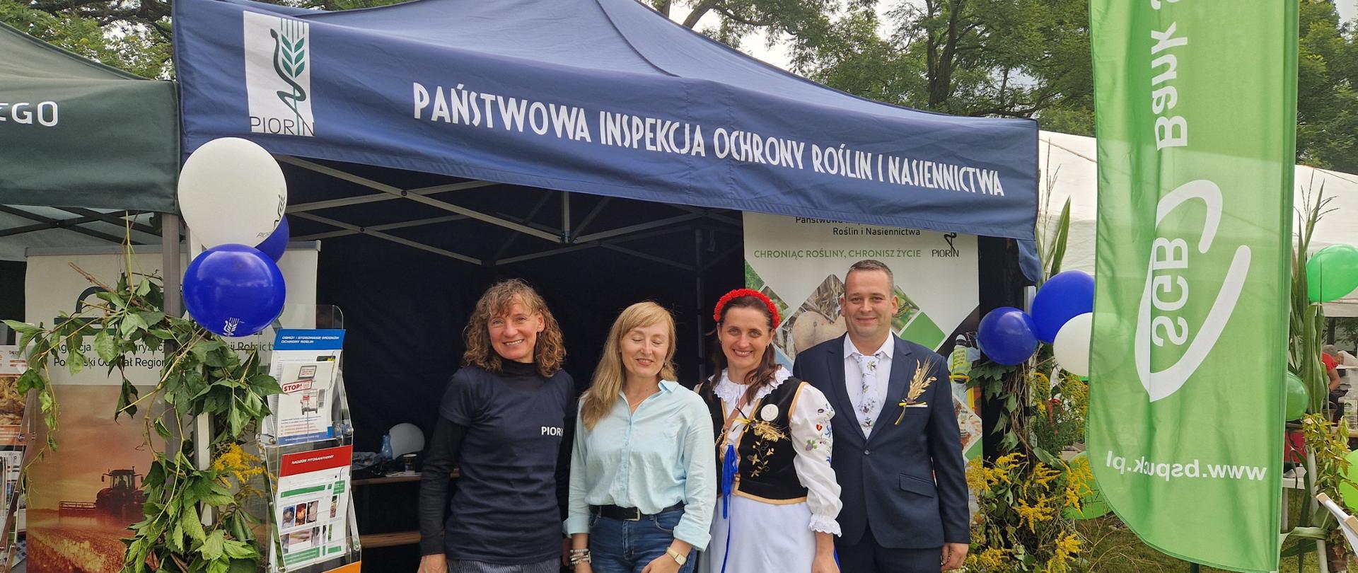 Cztery osoby, w tym kobieta w stroju ludowym, stoją przed namiotem Państwowej Inspekcji