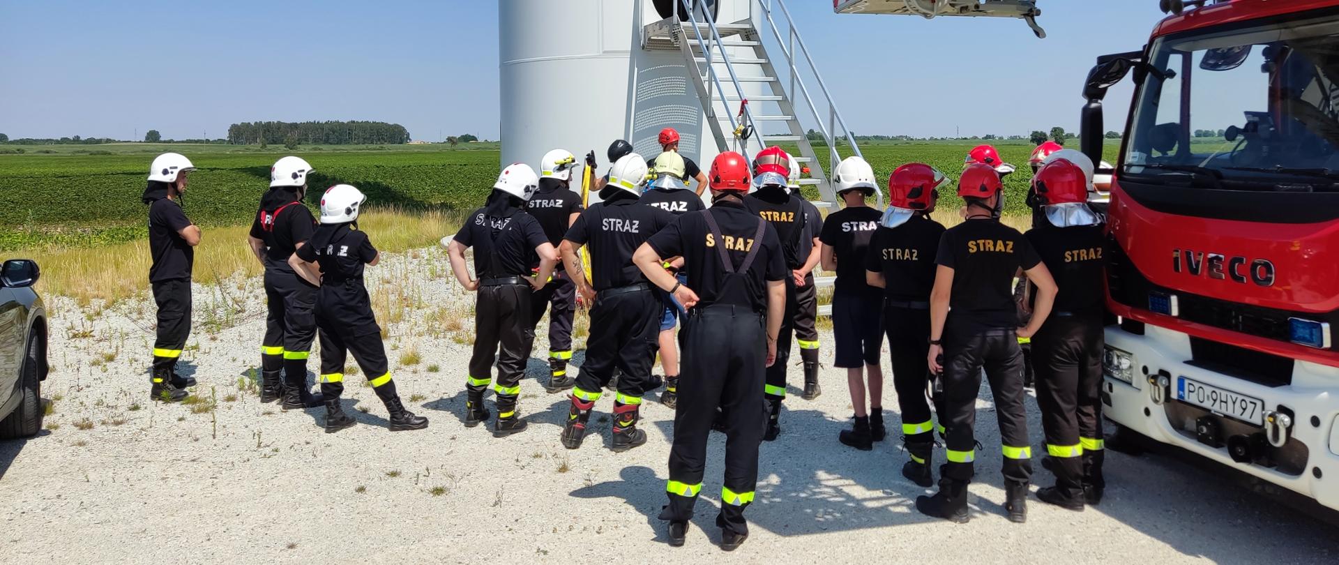 Zdjęcie przedstawia grupę strażaków stojących pod turbiną wiatrową, obok stoi podnośnik