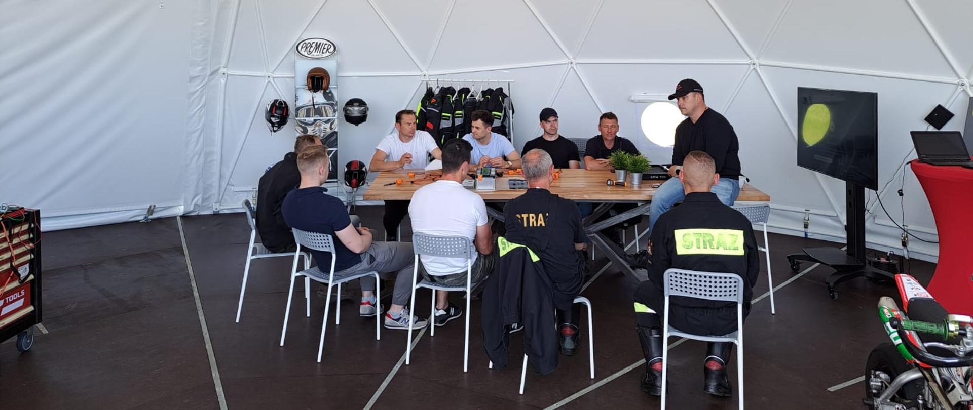 Na zdjęciu strażacy podczas szkolenia siedzący wokół podłużnego stołu, gdzie prezentowane są części baterii pojazdów elektrycznych