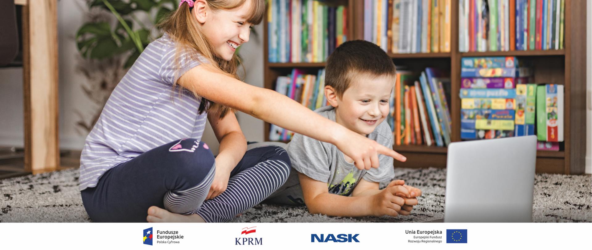 Na zdjęciu kilkuletnie dzieci - dziewczynka i chłopiec siedzą na dywanie i spoglądają w leżącego obok laptopa. Są uśmiechnięci, w tle regał z kolorowymi książkami i grami. 