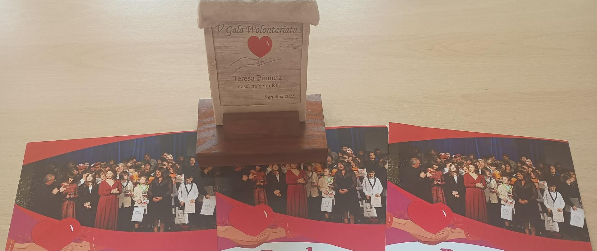 Na jasnym stole leżą trzy kolorowe dyplomy oraz drewniana tabliczka pamiątkowa. Każdy dyplom zawiera zdjęcie grupowe osób stojących na scenie podczas uroczystości. Dyplomy zostały przyznane trzem organizacjom szkolnym z ZSCKR w Oleszycach: Szkolnemu Klubowi Europejskiemu, Szkolnemu Kołu Caritas oraz Samorządowi Szkolnemu. Wszystkie wyróżnienia dotyczą działalności wolontariackiej, promowania idei pomocy i postaw humanitarnych. Nad dyplomami znajduje się drewniana plakietka z wygrawerowanym napisem „V Gala Wolontariatu”, imieniem i nazwiskiem Teresy Pamuły – Posłanki na Sejm RP – oraz datą: 8 grudnia 2025