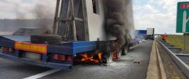 Ogień obejmuje koła i podwozie naczepy ciężarowej z ładunkiem płyt betonowych. Nad płonącym pojazdem unosi się gęsty, czarny dym.