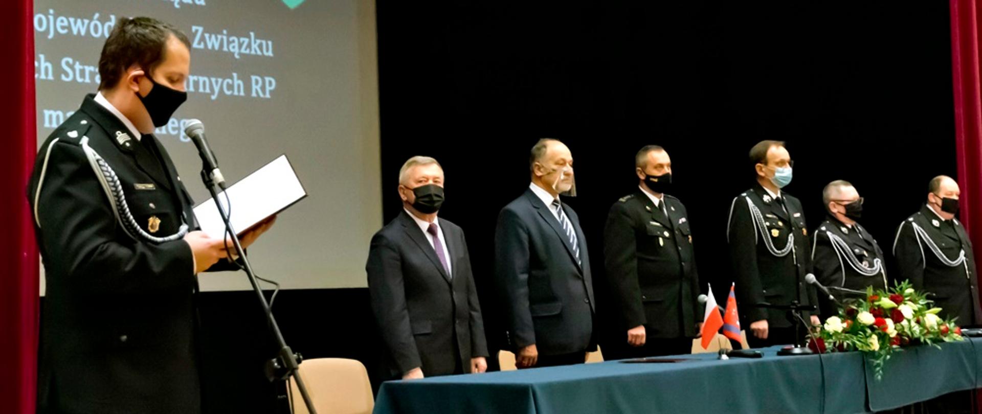 Osoby uczestniczące w posiedzeniu Zarządu OSP. Wszyscy ubrani na galowo. Z lewej strony jedna osoba stoi przy mikrofonie i odczytuje przemówienie.
