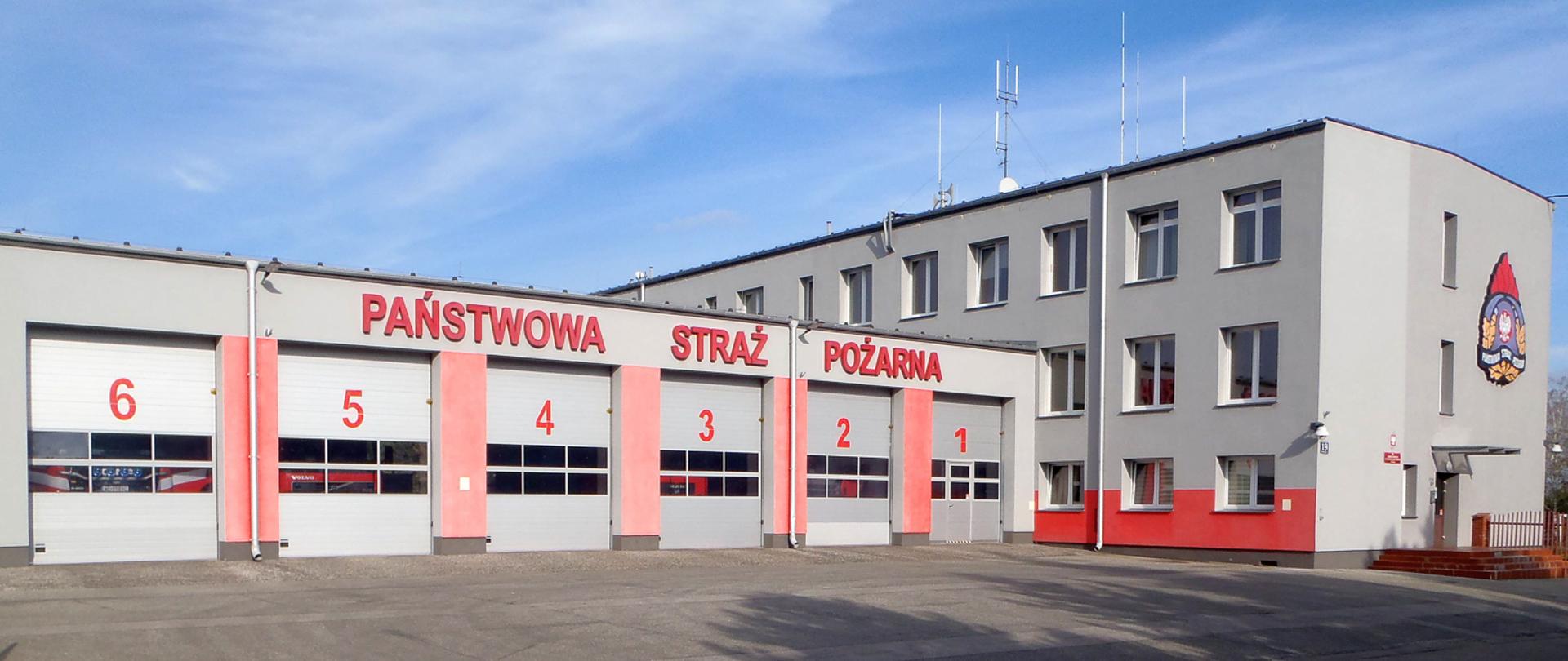 Zdjęcie przedstawia budynek Komendy Powiatowej PSP w Piasecznie w kolorze szaro-czerwonym. Na pierwszym planie bramy garażowe ponumerowane od 1 do 6. Z prawej strony zdjęcia wejście do siedziby piaseczyńskich strażaków.
