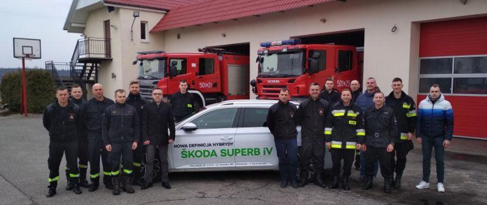 Zdjęcie zbiorowe strażaków na placu KP PSP na tle samochodu Skoda Superb iV i samochodów strażackich.