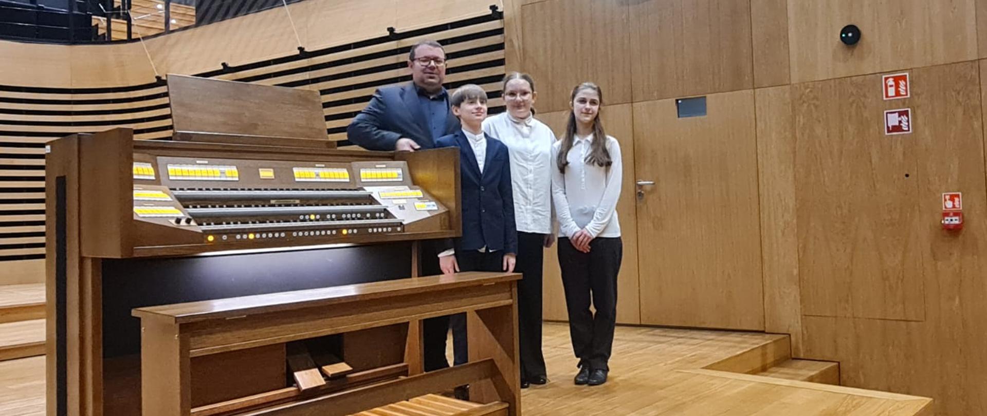 Uczniowie z nauczycielem stoją przy organach na estradzie.