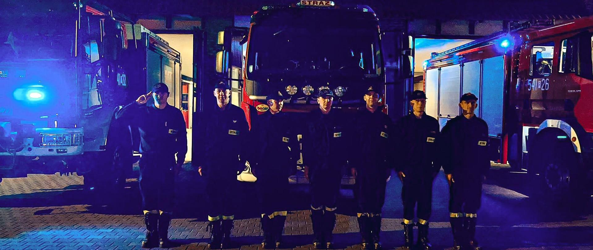 Strażacy stoją przed wozami strażackimi na placu manewrowym. Na drugim planie wozy strażackie i budynek strażnicy.