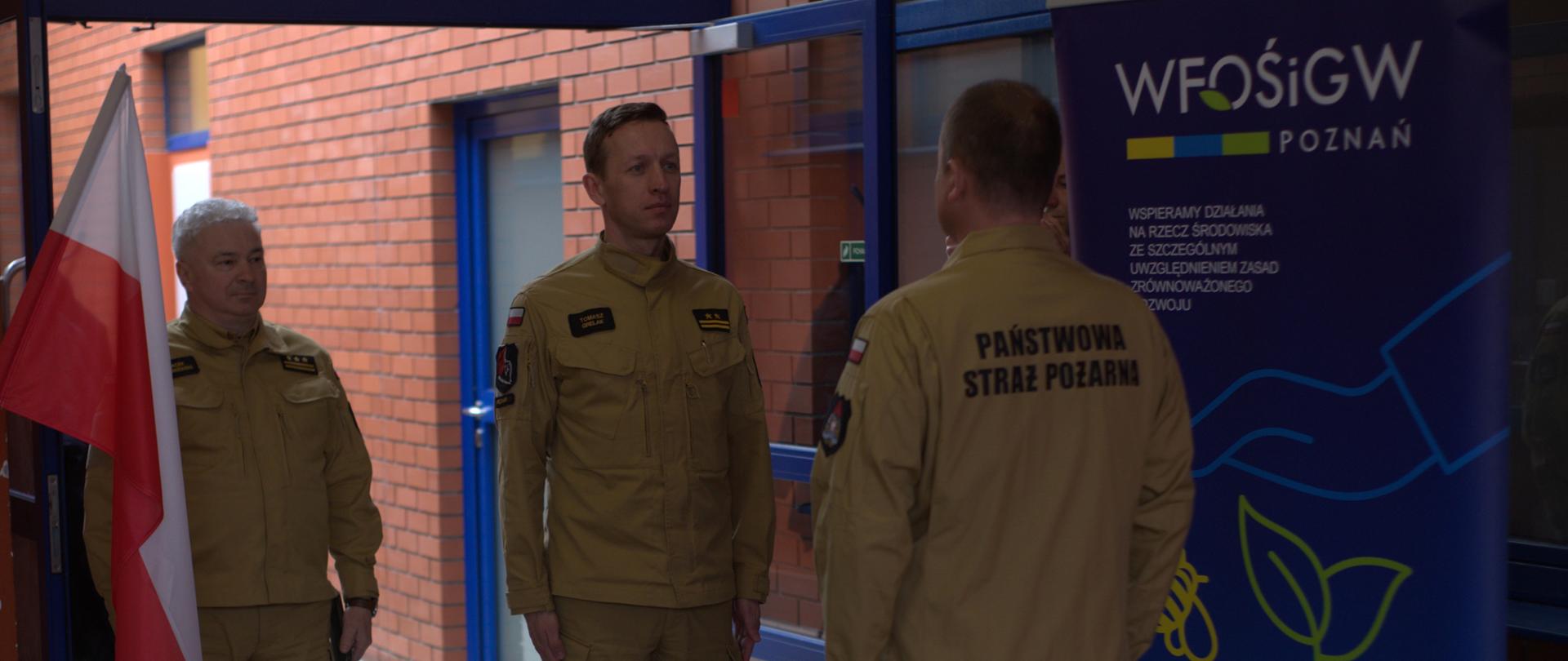 skłądanie meldunku strażakowi przez strażaka, w tle jeden strażak stoi na baczność oraz mury budynku