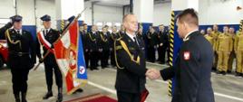Zdjęcie przedstawia zdającego obowiązki Komendanta Powiatowego odbierającego decyzje o zwolnieniu ze służby z rąk Zastępcy Komendanta Wojewódzkiego PSP. Za nimi poczet sztandarowy oddaje honory poprzez pochylenie sztandaru KP. Wokół zgromadzeni pracownicy i funkcjonariusze KP w Brodnicy.