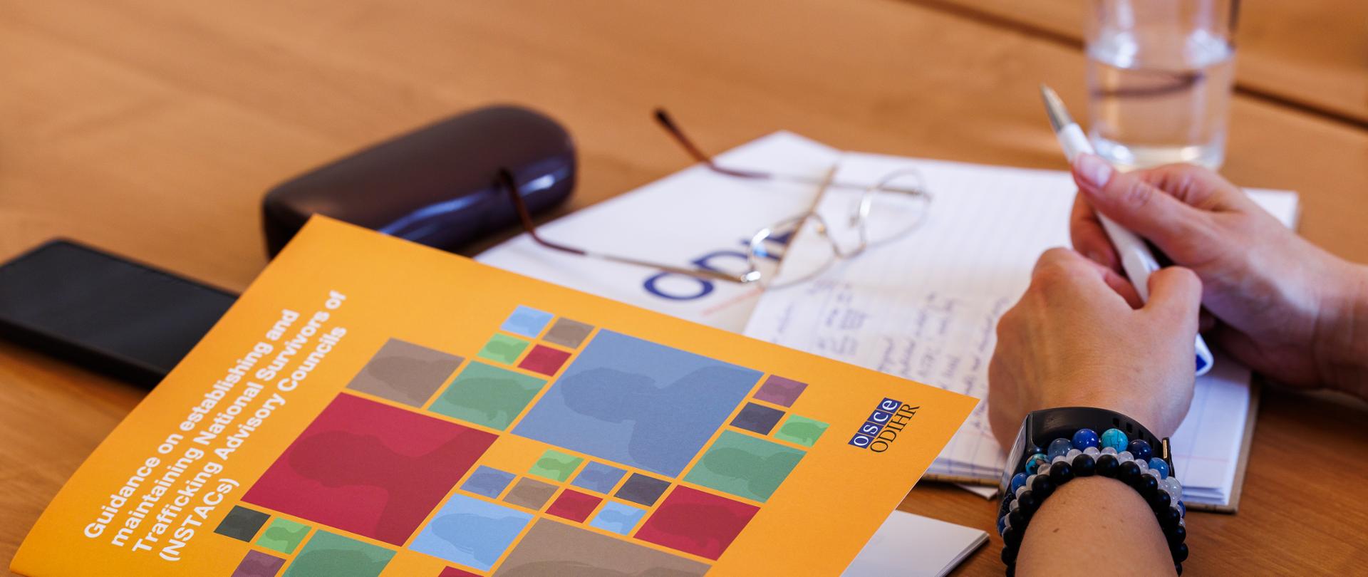 Na stole leżą okulary, zeszyt i Guidance on establishing and maintaining National Survivors of Trafficking Advisory Councils (NSTACs)