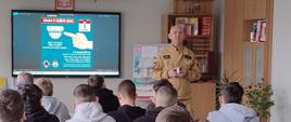 W sali lekcyjnej kilkunastu uczniów siedzi w ławkach, zwróconych w stronę nauczyciela w mundurze strażackim, który stoi przy tablicy multimedialnej. Na ekranie wyświetlona jest grafika dotycząca bezpieczeństwa pożarowego. W pomieszczeniu znajdują się regały z pomocami dydaktycznymi i rośliny.