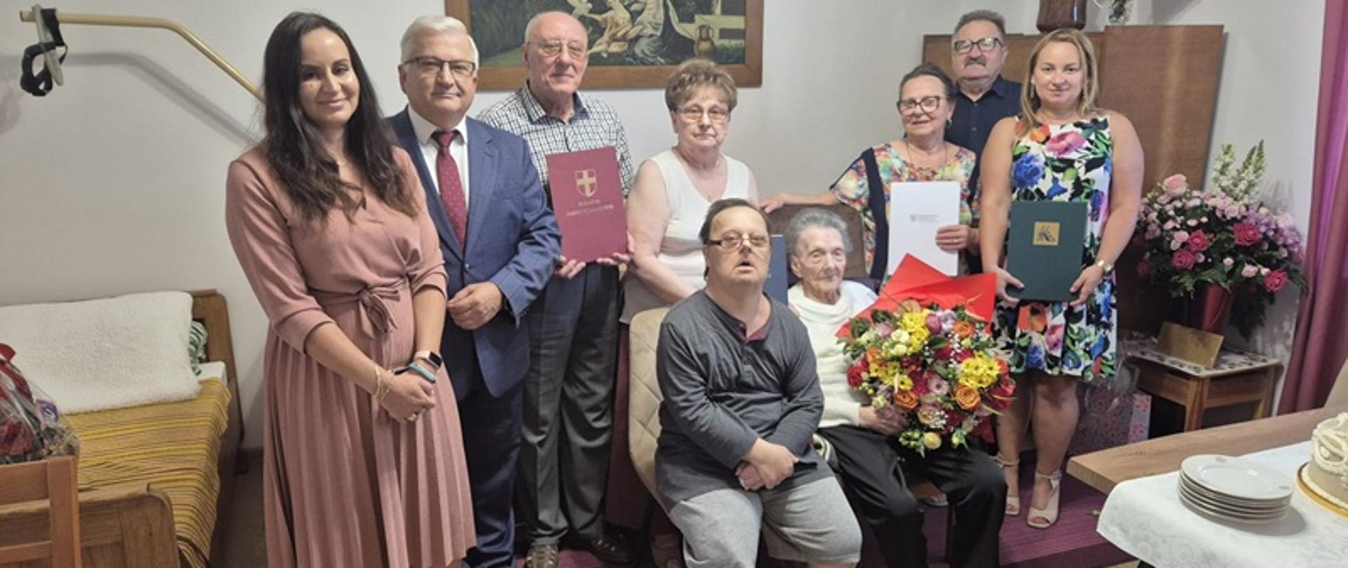 Na zdjęciu widać grupę dziewięciu osób zgromadzonych w pokoju. W centrum siedzi starsza kobieta trzymająca duży bukiet kwiatów; obok niej siedzi mężczyzna w okularach. Za nimi stoi sześć osób – cztery kobiety i dwóch mężczyzn, ubrani odświętnie, część z nich trzyma dokumenty w ozdobnych teczkach. Z lewej strony zdjęcia stoi kobieta w różowej sukience, z prawej kobieta w kwiecistej sukience. W tle widoczne są kwiaty w wazonach, obraz na ścianie i meble. Na pierwszym planie po prawej stronie znajduje się stół z białym obrusem i talerzykami, na którym stoi tort