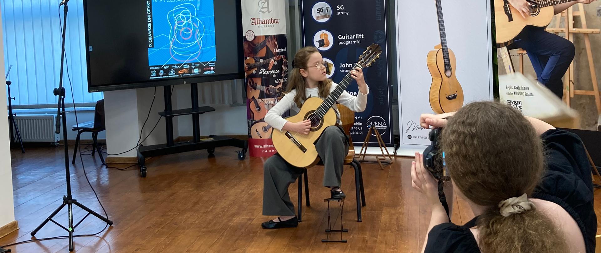 Sala koncertowa Orawskiej Samorządowej Szkoły Muzycznej w Jabłonce w dniu 19 maja 2023 r. Scena sali koncertowej szkoły muzycznej - uczennica naszej szkoły Amelia Lewkowicz podczas koncertu laureatów konkursu IX Ogólnopolskiego Konkursu Gitarowego. Na scenie siedzi na krzesełku, gra na gitarze. Po lewej stronie duży monitor, na którym jest wyświetlony plakat konkursu gitarowego w niebieskim kolorze. Po prawej stronie stoją banery reklamowe. Widać również tył głowy osoby siedzącej na widowni w prawym dolnym rogu.