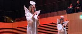Dwie kobiety w białych strojach aniołków przemawiają do mikrofonów, które trzymają w rękach.