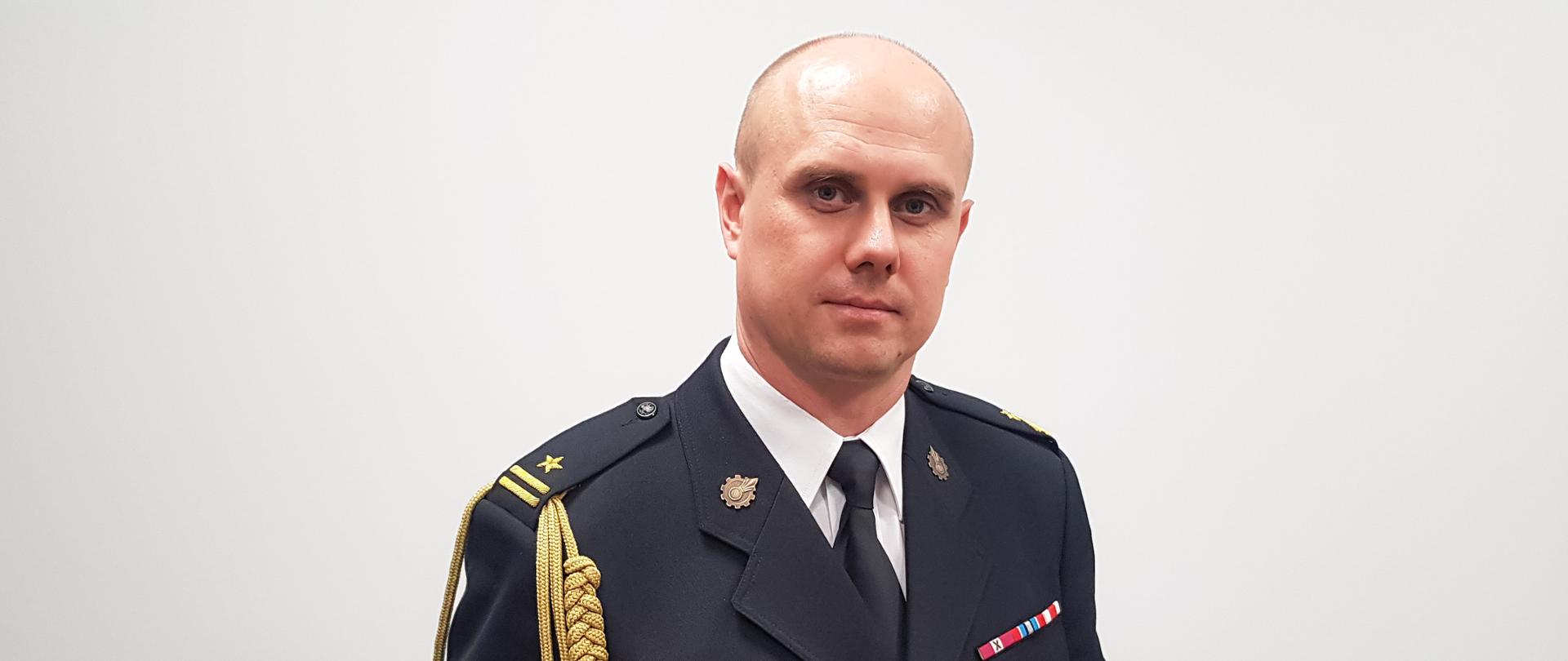 Zdjęcie portretowe komendanta mł.bryg. Łukasza Kapinosa na jasnym tle w mundurze galowym.