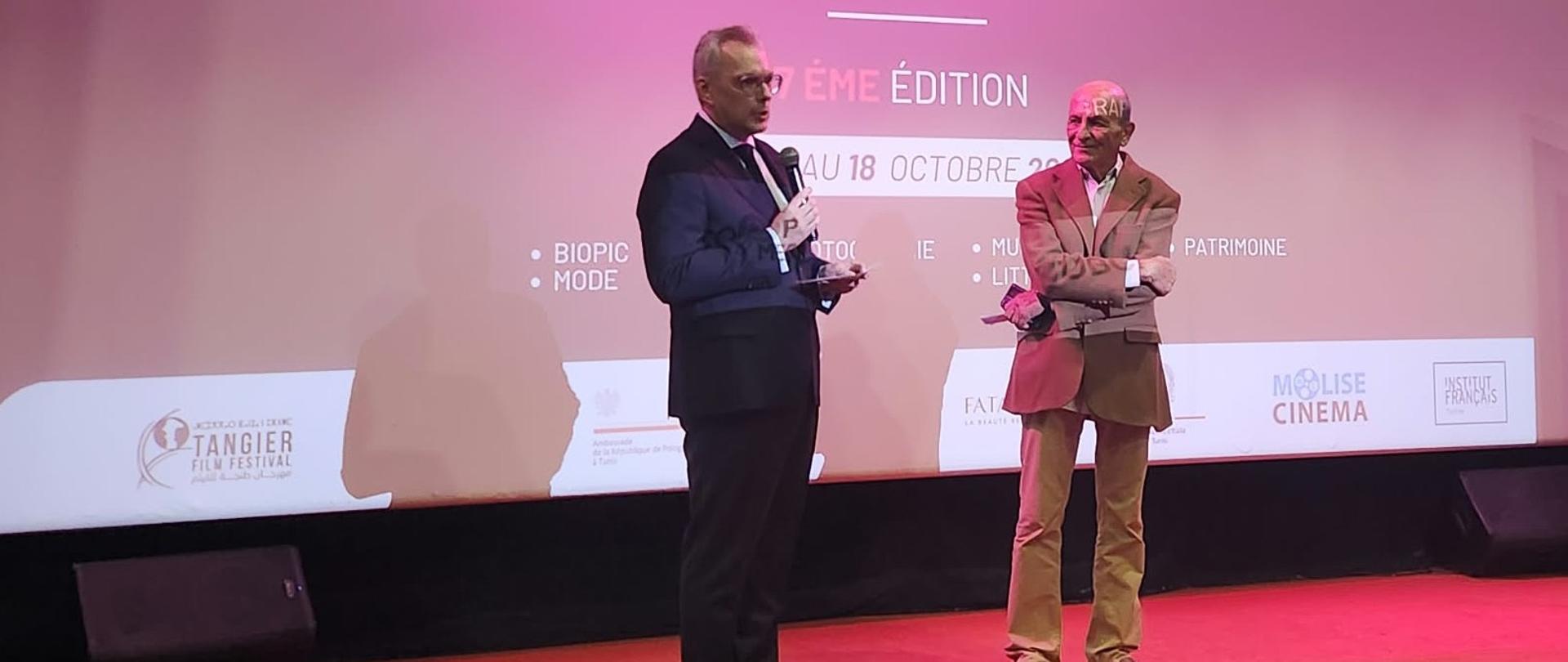 Na scenie kina Agora podczas festiwalu filmowego „Vues sur les Arts” Ambasador RP w Tunisie przemawia do publiczności, trzymając mikrofon. Obok niego stoi mężczyzna w beżowym garniturze. W tle widoczny jest ekran z nazwą festiwalu.