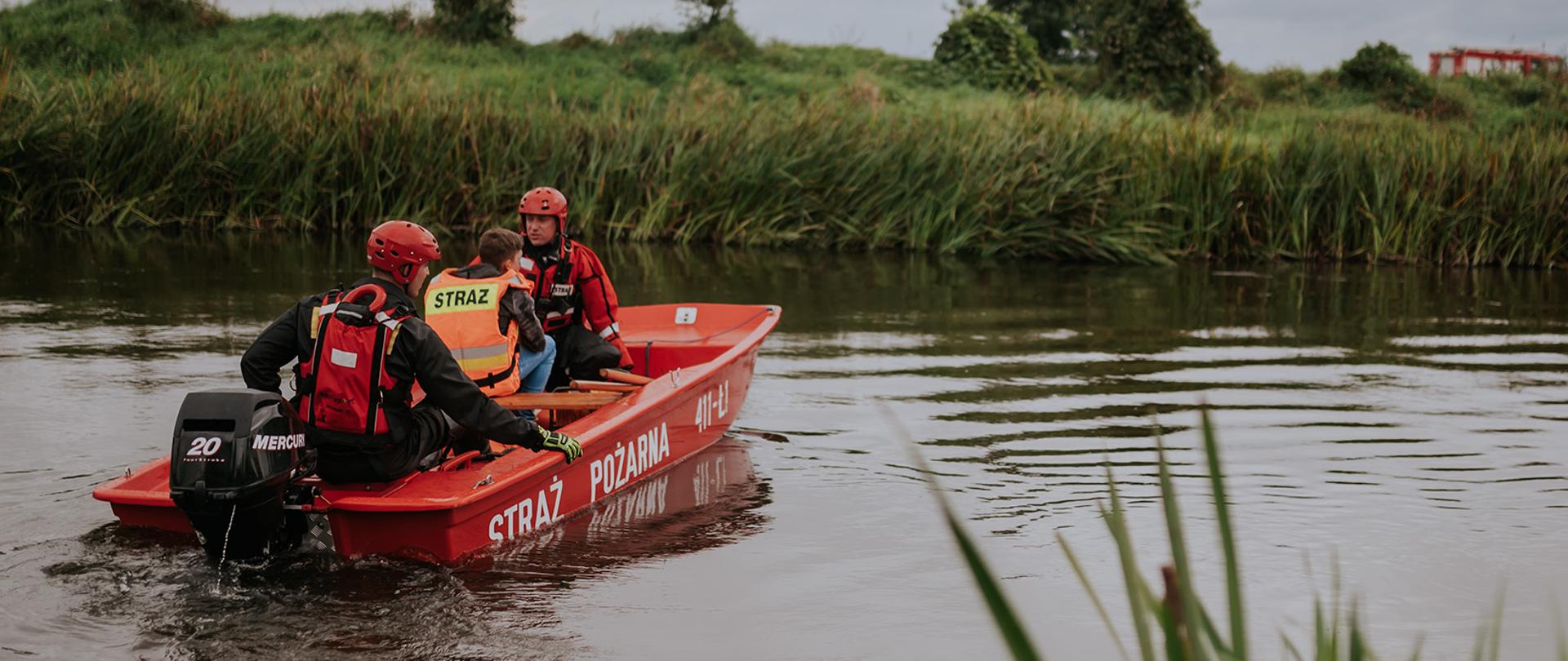 Na łodzi płaskodennej dwóch strażaków przewozi poszkodowanego harcerza na drugi brzeg rzeki.