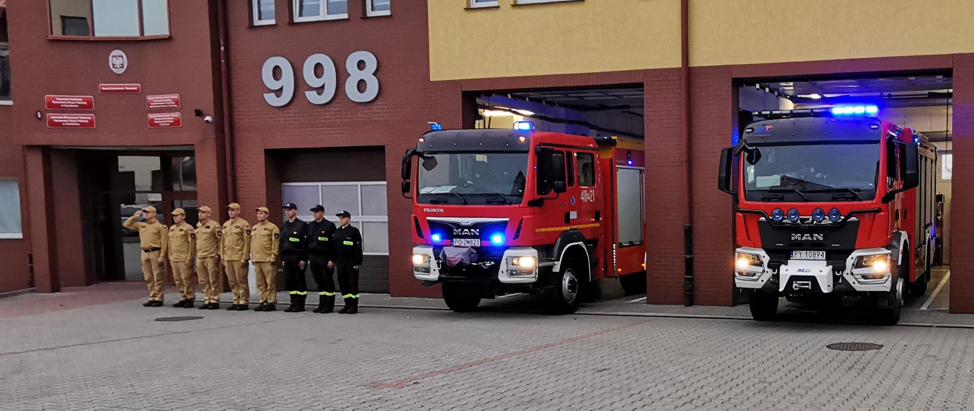 Strażacy w umundurowaniu stoją w szeregu przed budynkiem Komendy Powiatowej PSP w Czarnkowie, na placu ustawione są dwa wozy pożarnicze z włączonymi sygnałami świetlnymi.