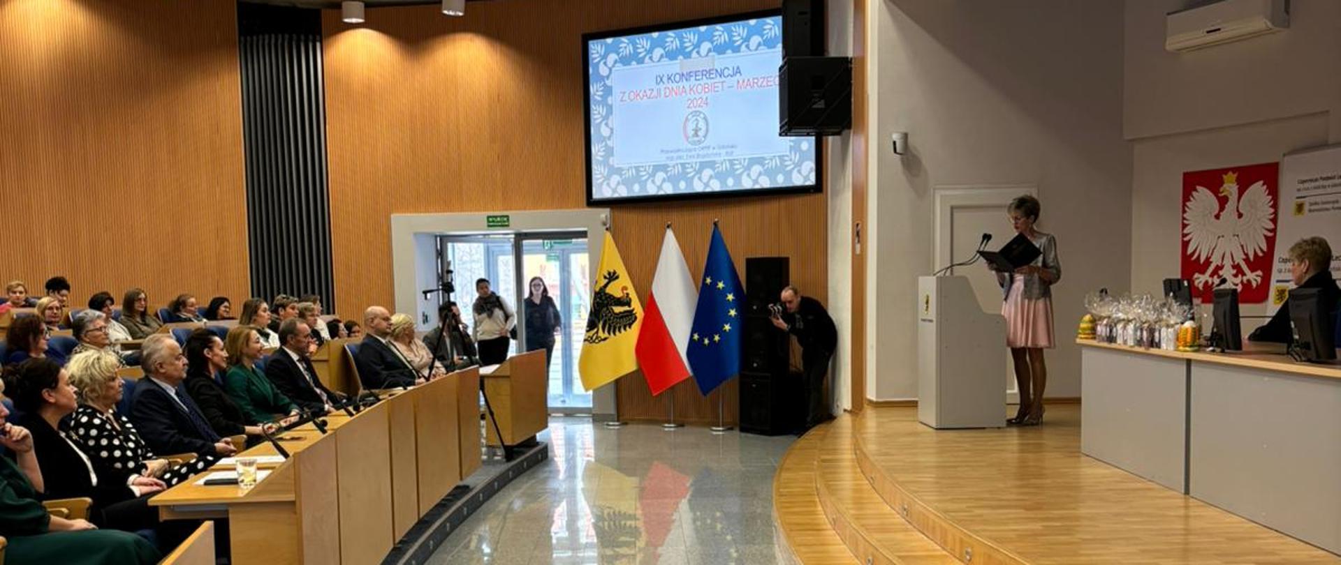 duża sala, goście siedzą w fotelach i słuchają wykładu, w tle tablica z napisem Konferencja z okazji Dnia Kobiet
