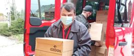 Na zdjęciu widoczni strażacy podczas rozładunku płynu do dezynfekcji i maseczek. Na pierwszym planie strażak ochotnik z kartonem w rękach w ubraniu cywilnym czarna kurtka czarne spodnie na drugim planie strażak PSP w ubraniu dowódczo sztabowym biorący karton z czerwonego samochodu pożarniczego. 