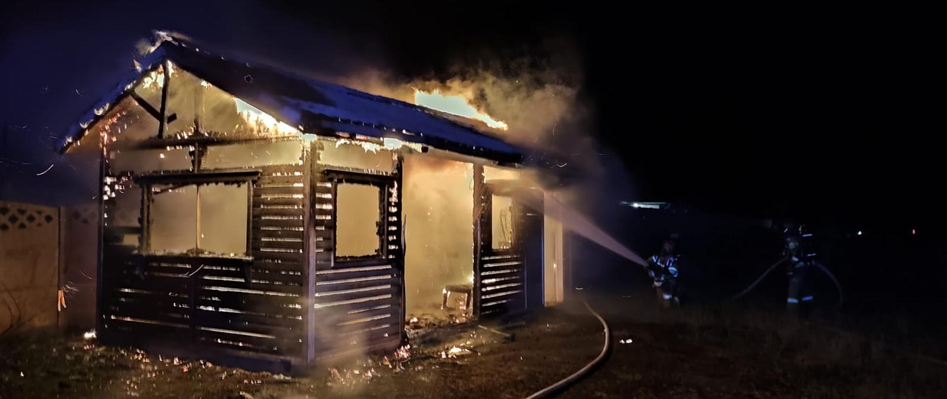 Na zdjęciu paląca się altana oraz strażacy gaszący obiekt w aparatach ochrony dróg oddechowych