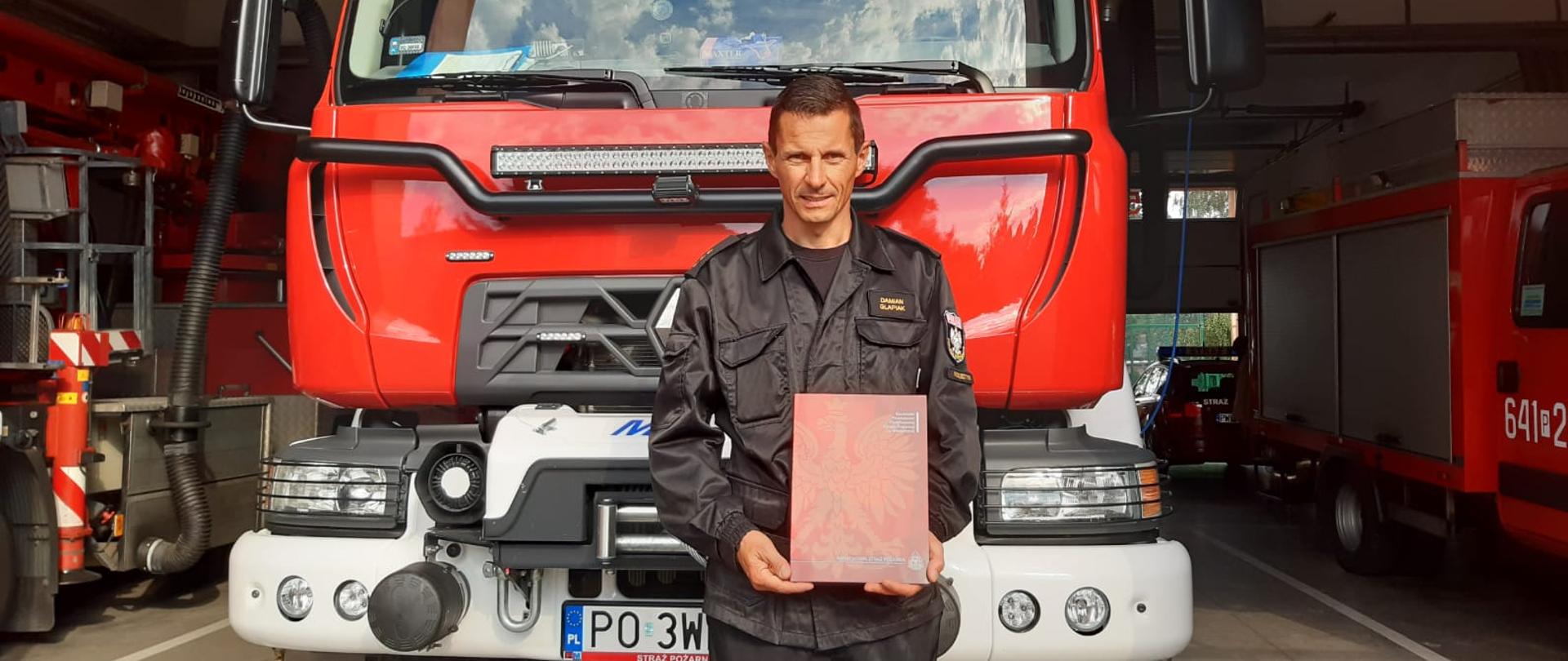 Funkcjonariusz stoi na tle samochodu gaśniczego. W rękach trzyma teczkę.