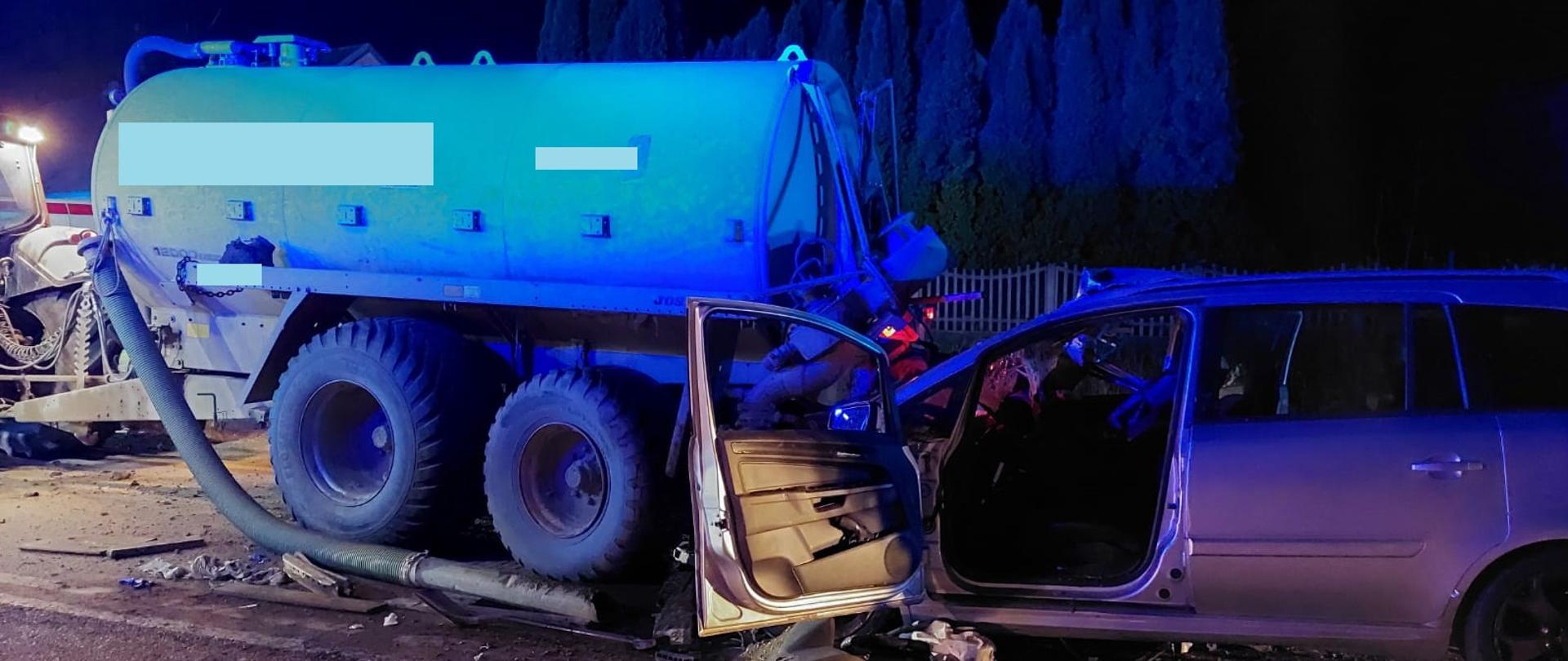 Na zdjęciu widoczny jest wrak samochodu osobowego oraz zestaw ciągnika rolniczego z beczką. 