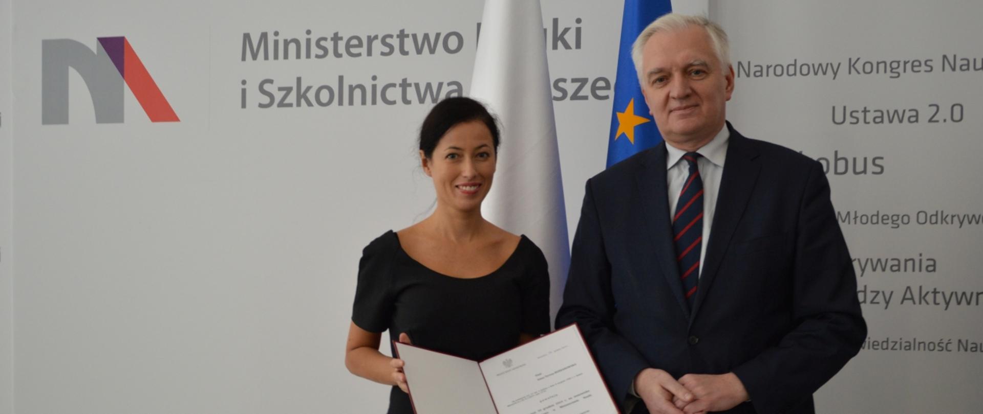 Dr Anna Budzanowska i minister Jarosław Gowin stoją na tle ściany. Dr Budzanowska trzyma powołanie na funkcję podsekretarza stanu.