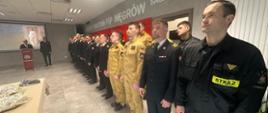 Na zdjęciu widoczna jest uroczystość odbywająca się w siedzibie Komendy Powiatowej Państwowej Straży Pożarnej. Wzdłuż sali ustawieni są strażacy w umundurowaniu wyjściowym oraz służbowym, stojący w szeregu podczas oficjalnego wystąpienia. Po lewej stronie znajduje się podium, przy którym przemawia osoba prowadząca uroczystość. W tle widoczny jest ekran z wyświetlonym wizerunkiem o charakterze religijnym, . 