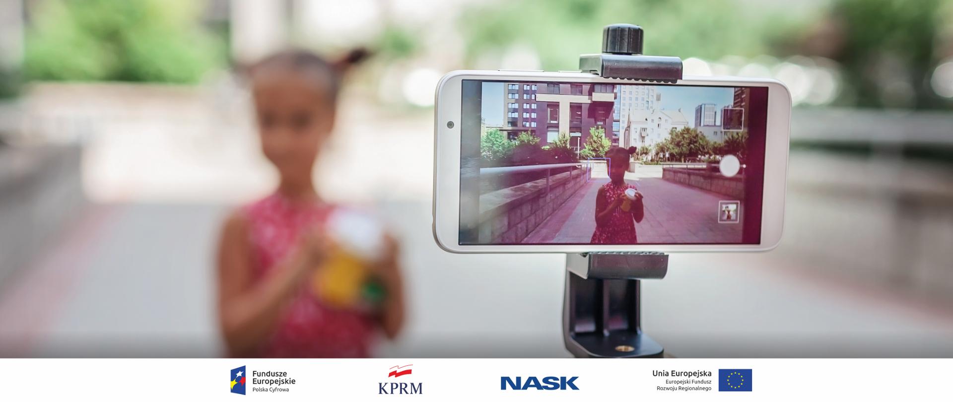 Zdjęcie przedstawiające smartfona na statywie z uruchomionym aparatem fotograficznym. Na uchwyconym kadrze widać dziewczynkę z kubkiem, w tle widać budynki. Na dalszym planie widać rozmyty obraz dziewczynki pozującej do zdjęcia. 