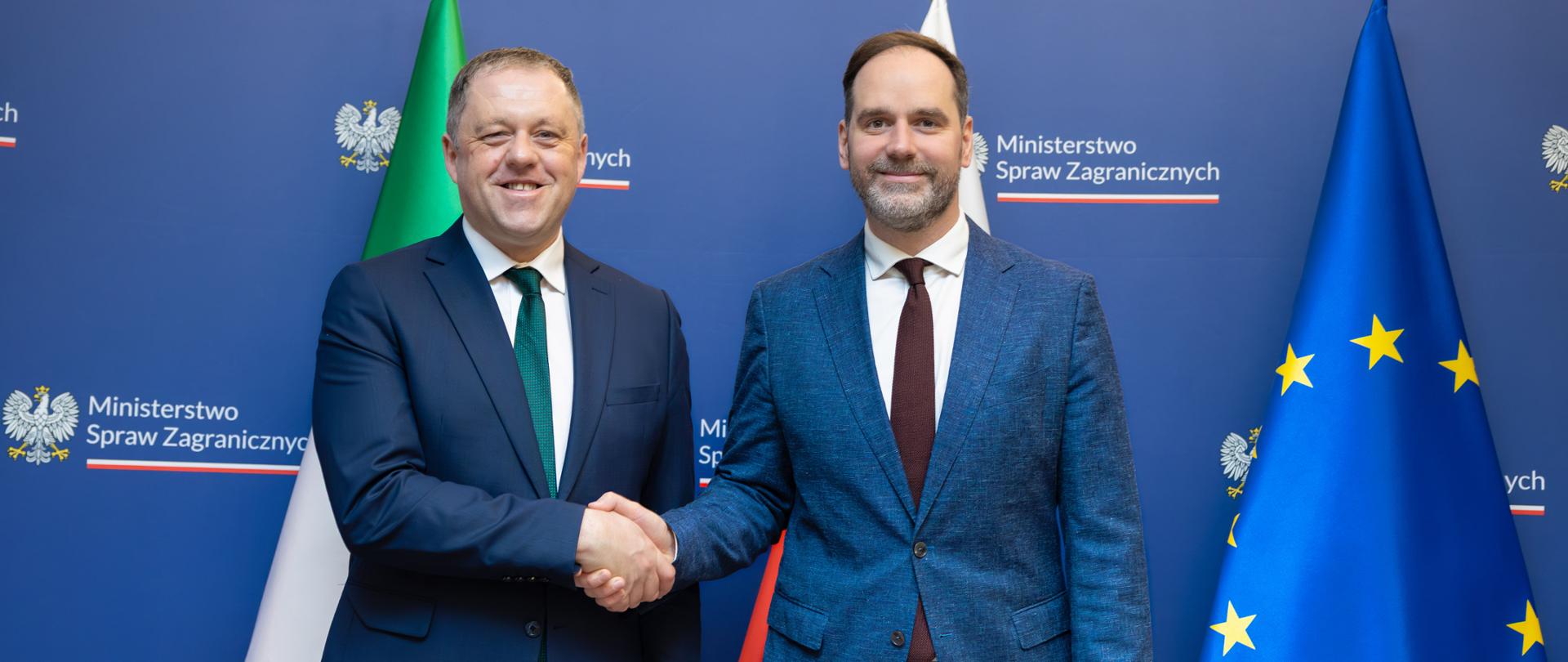 Na zdjęciu jest dwóch mężczyzn w garniturach. Stoją, podają sobie dłonie, są zwróceni w kierunku obiektywu. Za nimi są 3 flagi: Irlandii, Polski i Unii Europejskiej. 