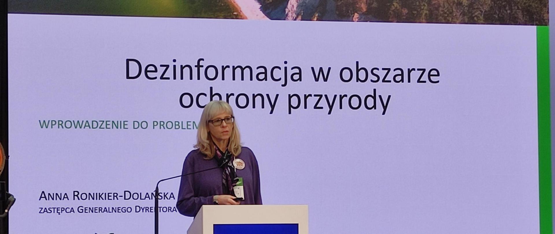 Na tle wyświetlonego slajdu z napisami: Dezinformacja w obszarze ochrony przyrody. Wprowadzenie do problemu. Anna Ronikier-Dolańska, Zastępca Generalnego Dyrektora, przy mównicy z mikrofonem stoi kobieta. Kobieta trzyma w ręku pilot zdalnego sterowania. 