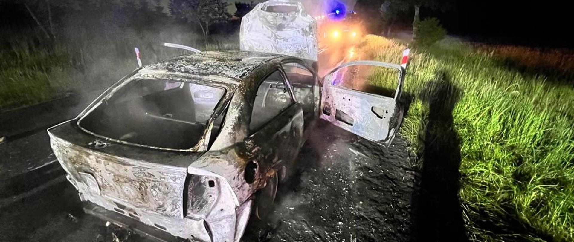 Zdjęcie przedstawia spalony wrak samochodu osobowego na poboczu drogi. 