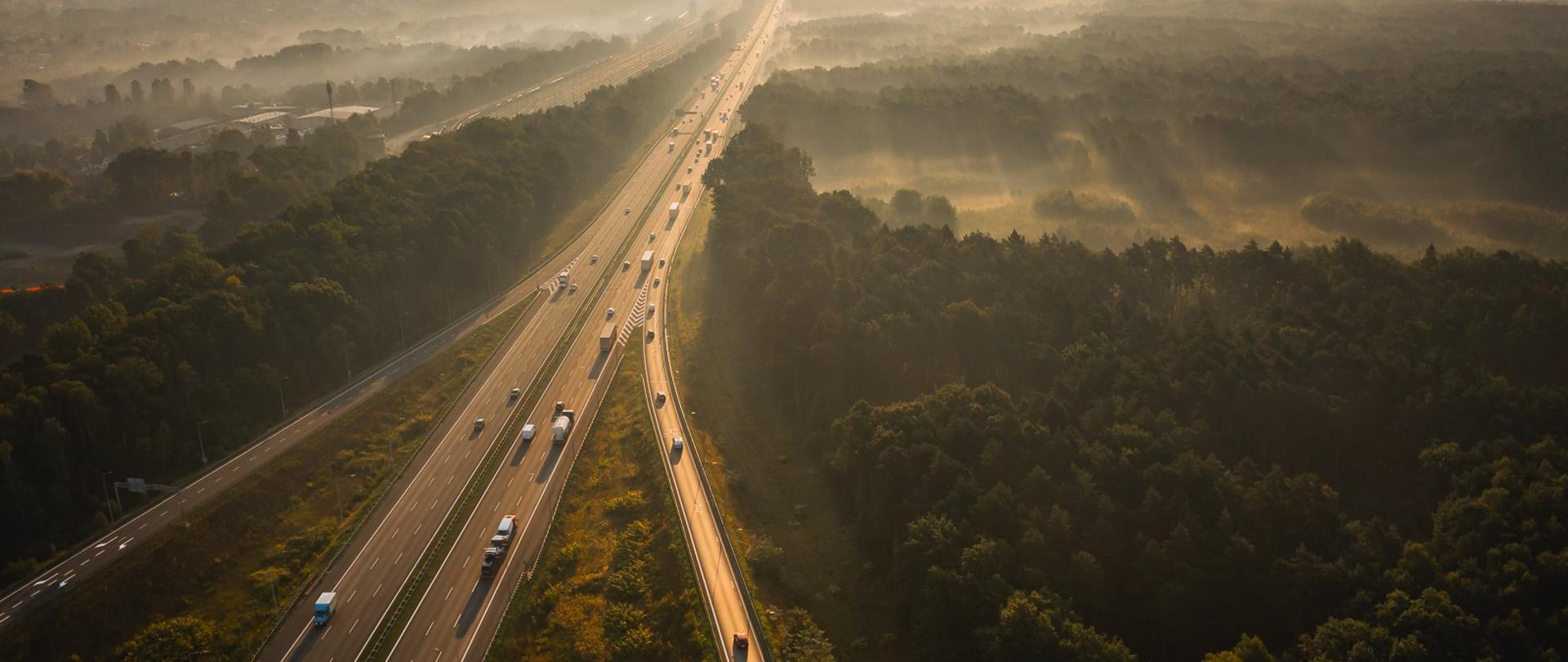 Zdjęcie przedstawia autostradę biegnącą przez zalesiony teren, widzianą z lotu ptaka. Na drodze widać kilka samochodów jadących w obu kierunkach. Po lewej stronie znajduje się dwujezdniowa szeroka autostrada, a po prawej stronie jednopasmowa jezdnia z której pojazdy włączają się do ruchu na autostradzie. Krajobraz jest oświetlony ciepłym światłem zachodzącego lub wschodzącego słońca, co nadaje zdjęciu malowniczy i spokojny charakter. W tle można dostrzec mgłę unoszącą się nad lasem