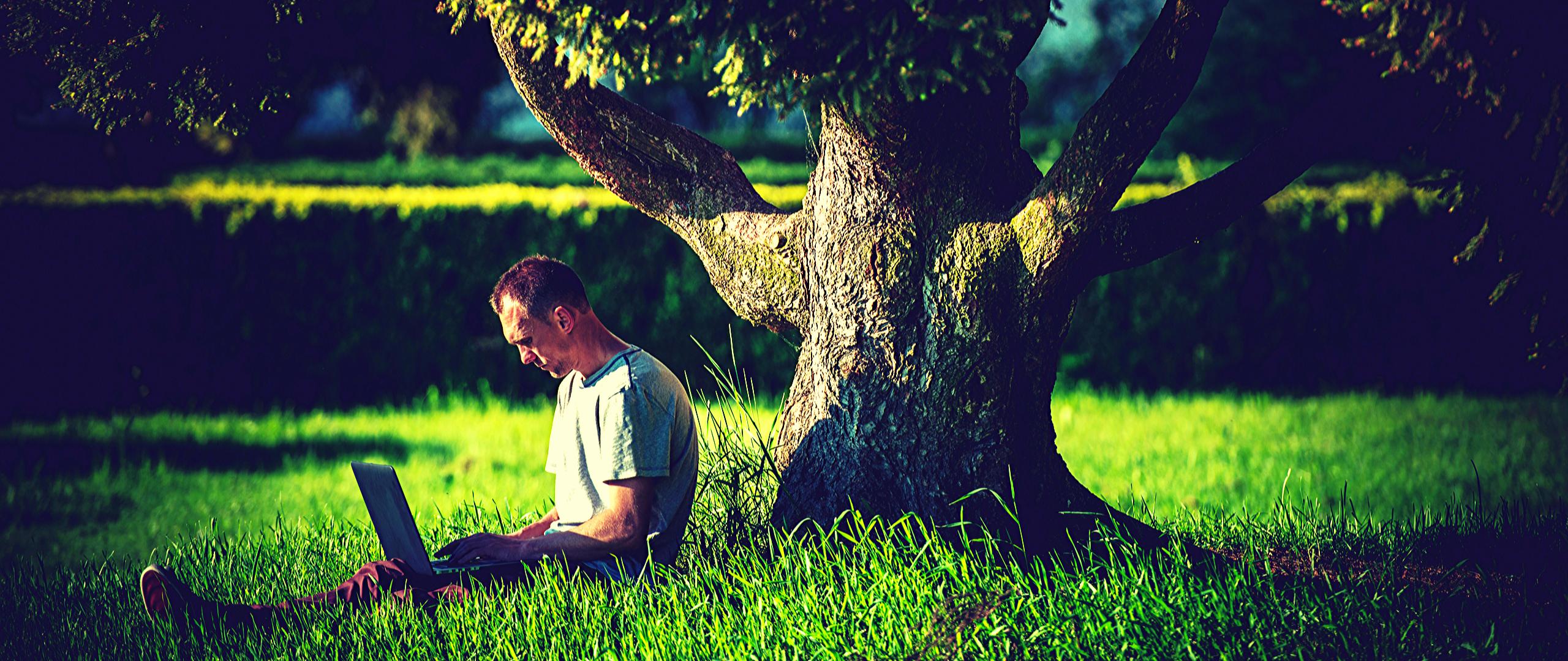Mężczyzna siedzący pod drzewem z laptopem na kolanach
