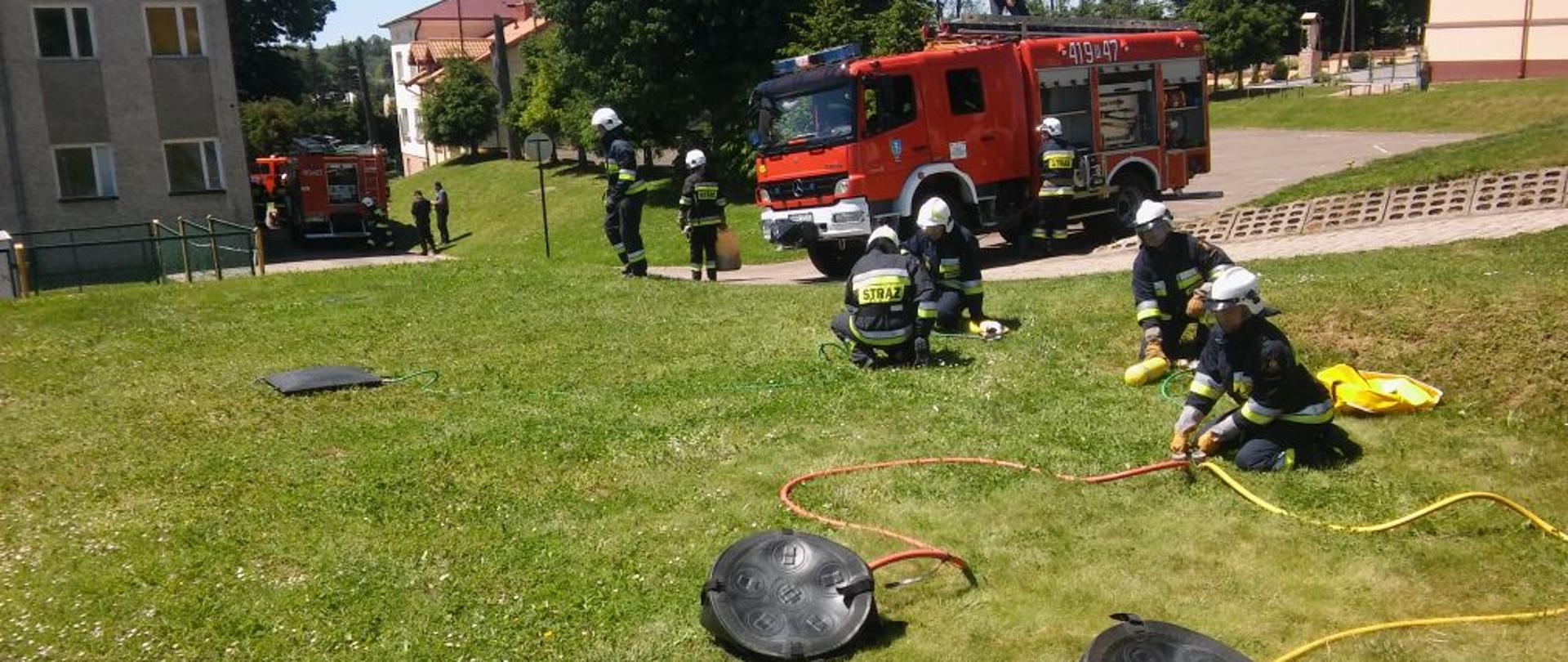 Przykładowe zdjęcie przedstawiające szkolenie Ochotniczych Straży Pożarnych. Na pierwszym planie zielona trawa, na której kilku umundurowanych druhów Ochotniczych Straży Pożarnych ćwiczy ze sprzętem pneumatycznym. Na drugim planie czerwony ciężarowy pojazd ratowniczo-gaśniczy, przy nim kilku druhów. W tle budynki, zielone drzewa i inny pojazd pożarniczy i kilku druhów.