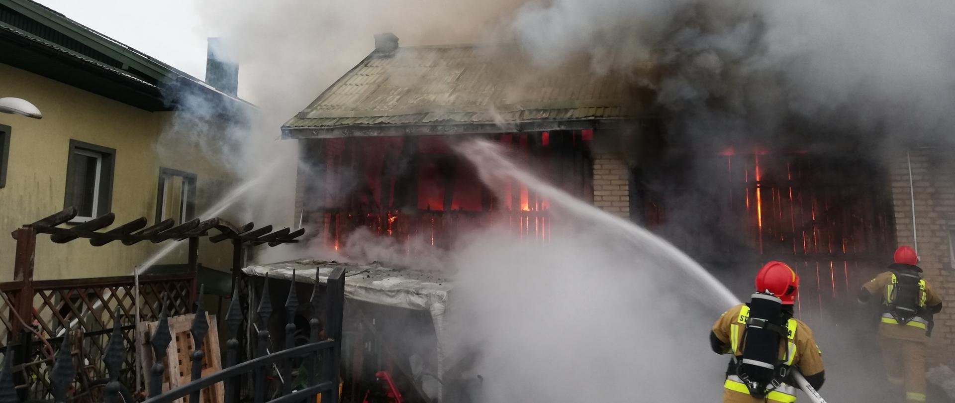 Płonący budynek gospodarczy, ogień wychodzi na zewnątrz, Strażacy podający wodę na pożar i w obronie budynku sąsiedniego 